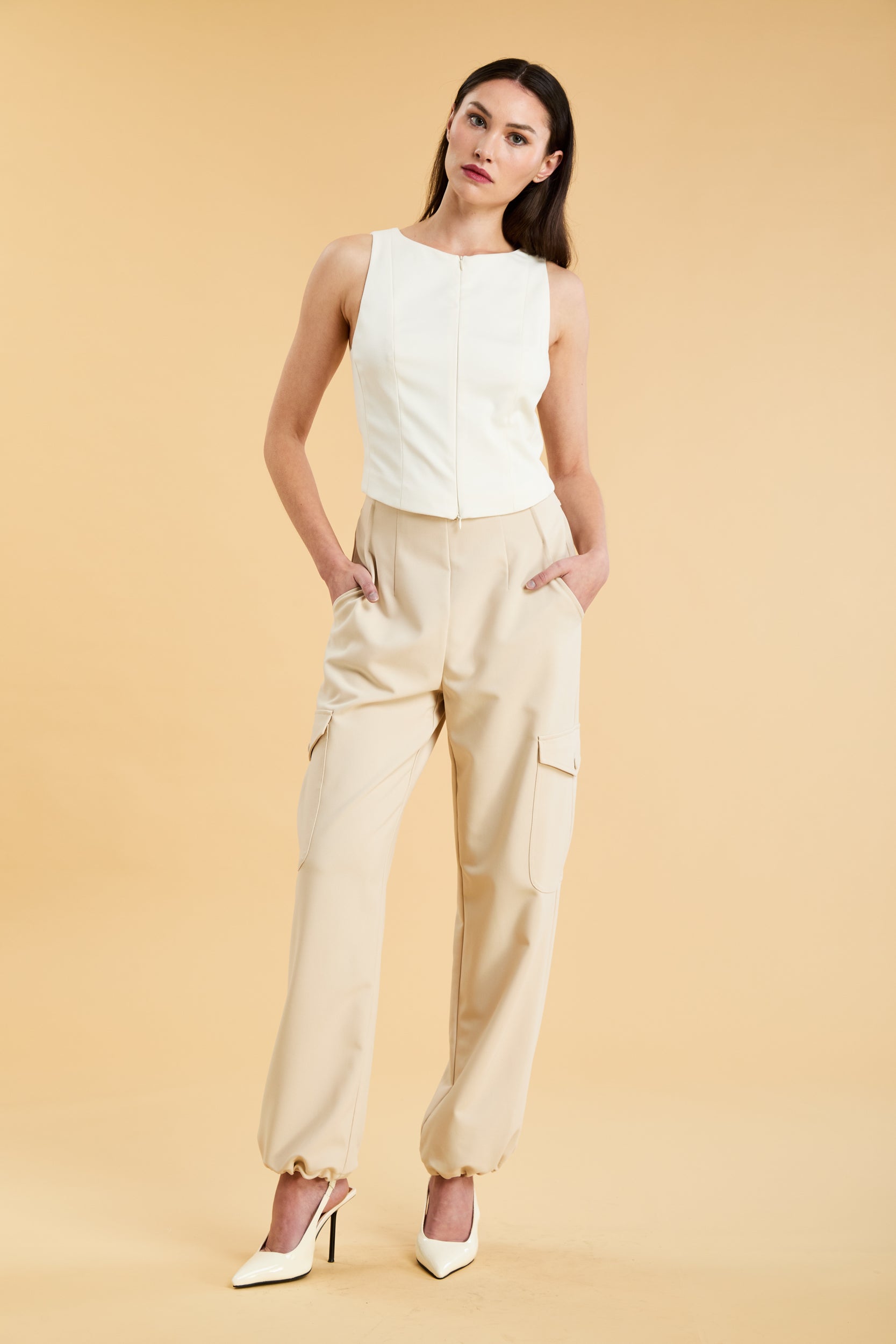 Woman wearing a white sleeveless double zipper top and beige almond Triplicity trousers cargo pants on a beige background