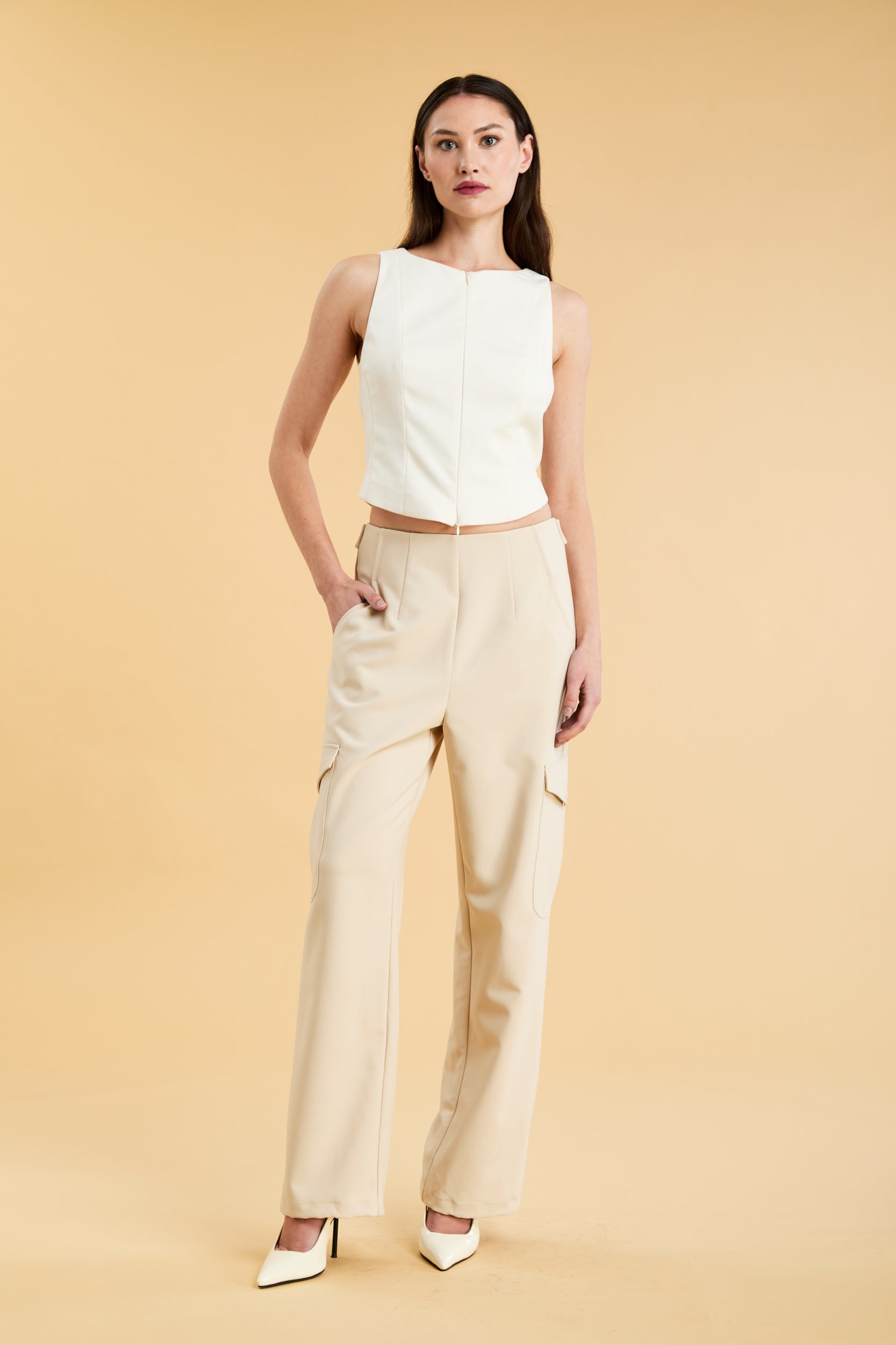 Woman wearing a white sleeveless double zipper top and beige almond Triplicity trousers cargo pants on a beige background