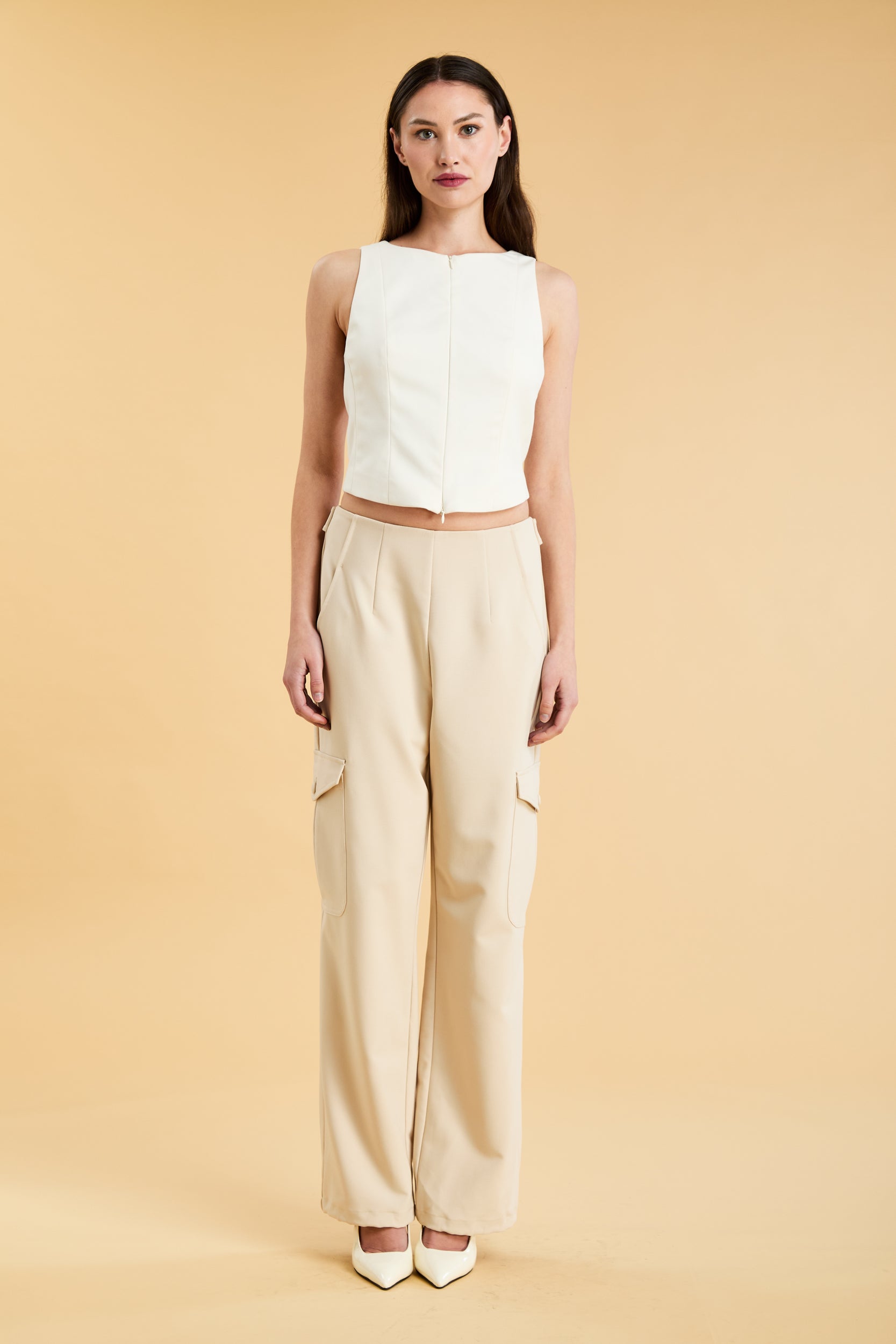Woman wearing a white sleeveless double zipper top and beige almond Triplicity trousers cargo pants on a beige background