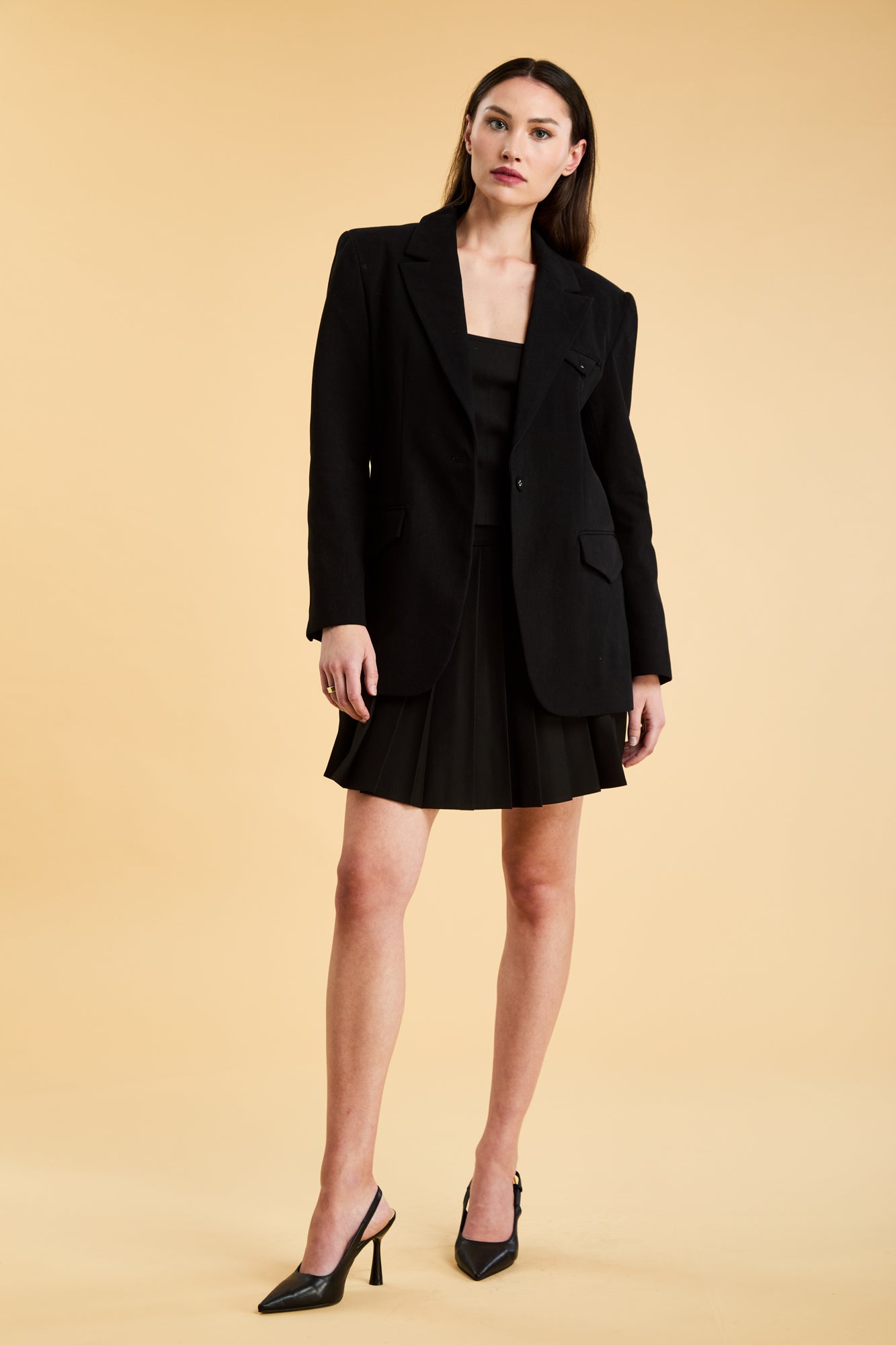 Woman wearing a Triplicity Black Constance Blazer jacket and a Black Pleated Elisabeth mini skirt on a beige background