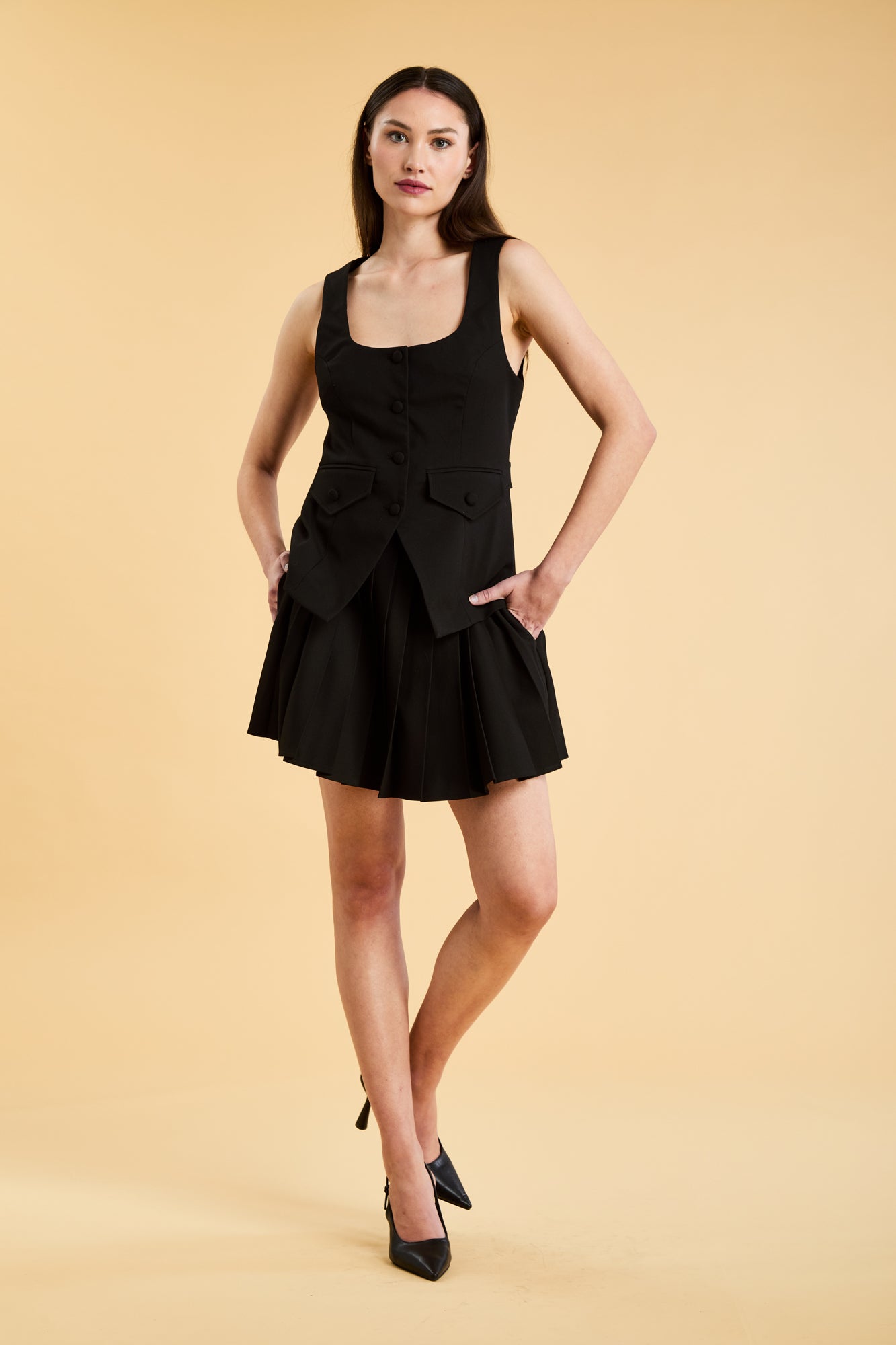 Woman wearing a Triplicity Black Georgiana Vest top and a Black Pleated Elisabeth mini skirt on a beige background