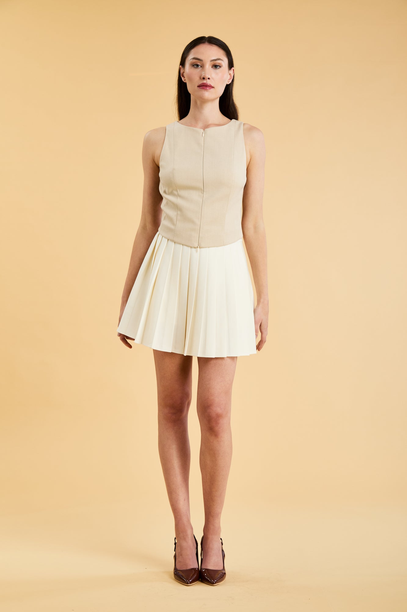 Woman wearing a Triplicity Alice Oat sleeveless double ended zipper top and Triplicity White pleated mini skirt against a beige background