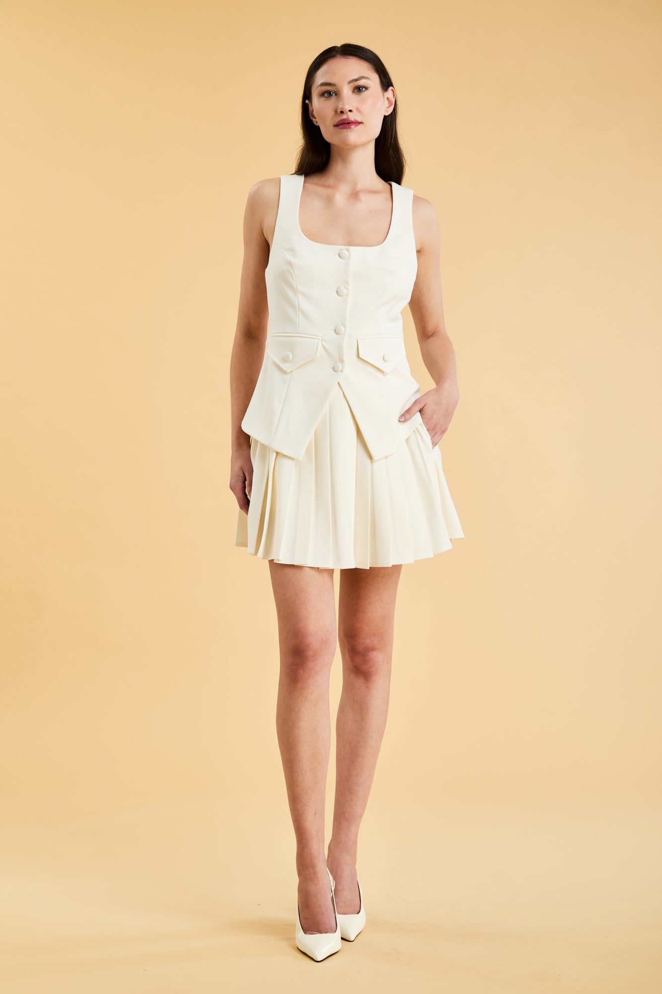 Woman wearing a Triplicity Georgiana White Vest Top and Triplicity White pleated mini skirt against a beige background full body front view