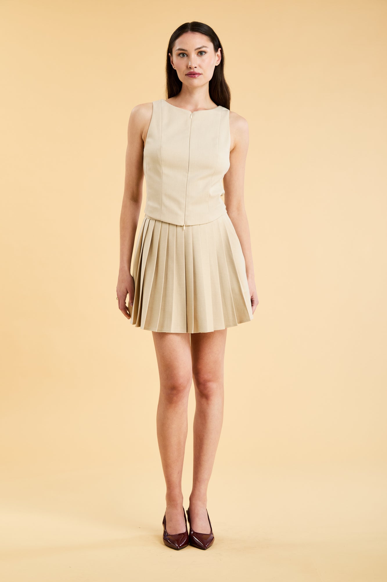 Woman wearing a Oat colored Triplicity Eloise Fitted Tank Top and a Oat Pleated Elisabeth mini skirt on a beige background full body front view