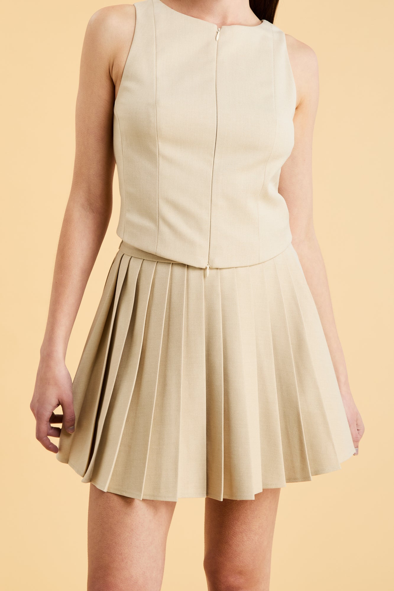 Woman wearing a Oat colored Triplicity Eloise Fitted Tank Top and a Oat Pleated Elisabeth mini skirt on a beige background close up front view