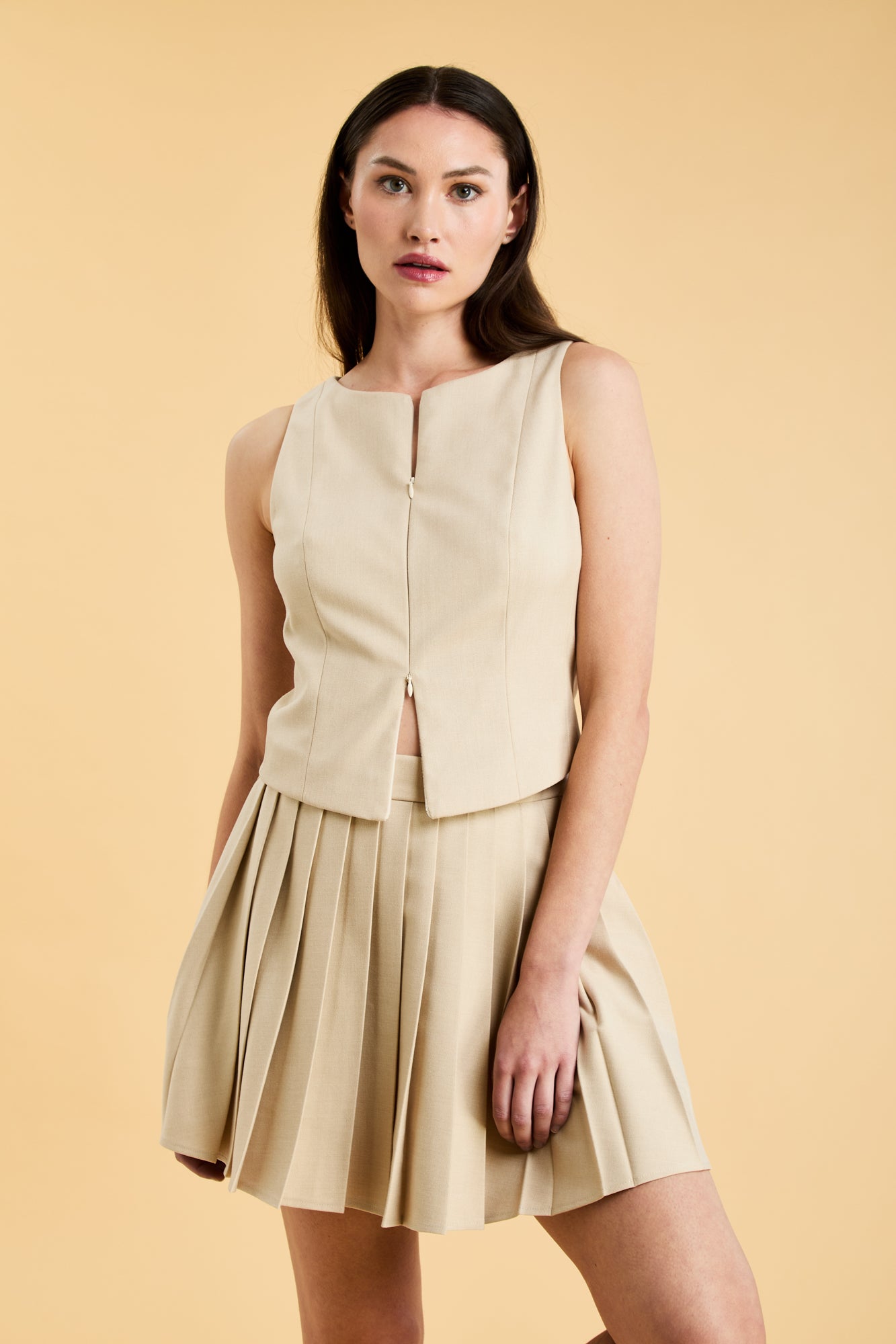 Woman wearing a Oat colored Triplicity Eloise Fitted Tank Top and a Oat Pleated Elisabeth mini skirt on a beige background close up front view