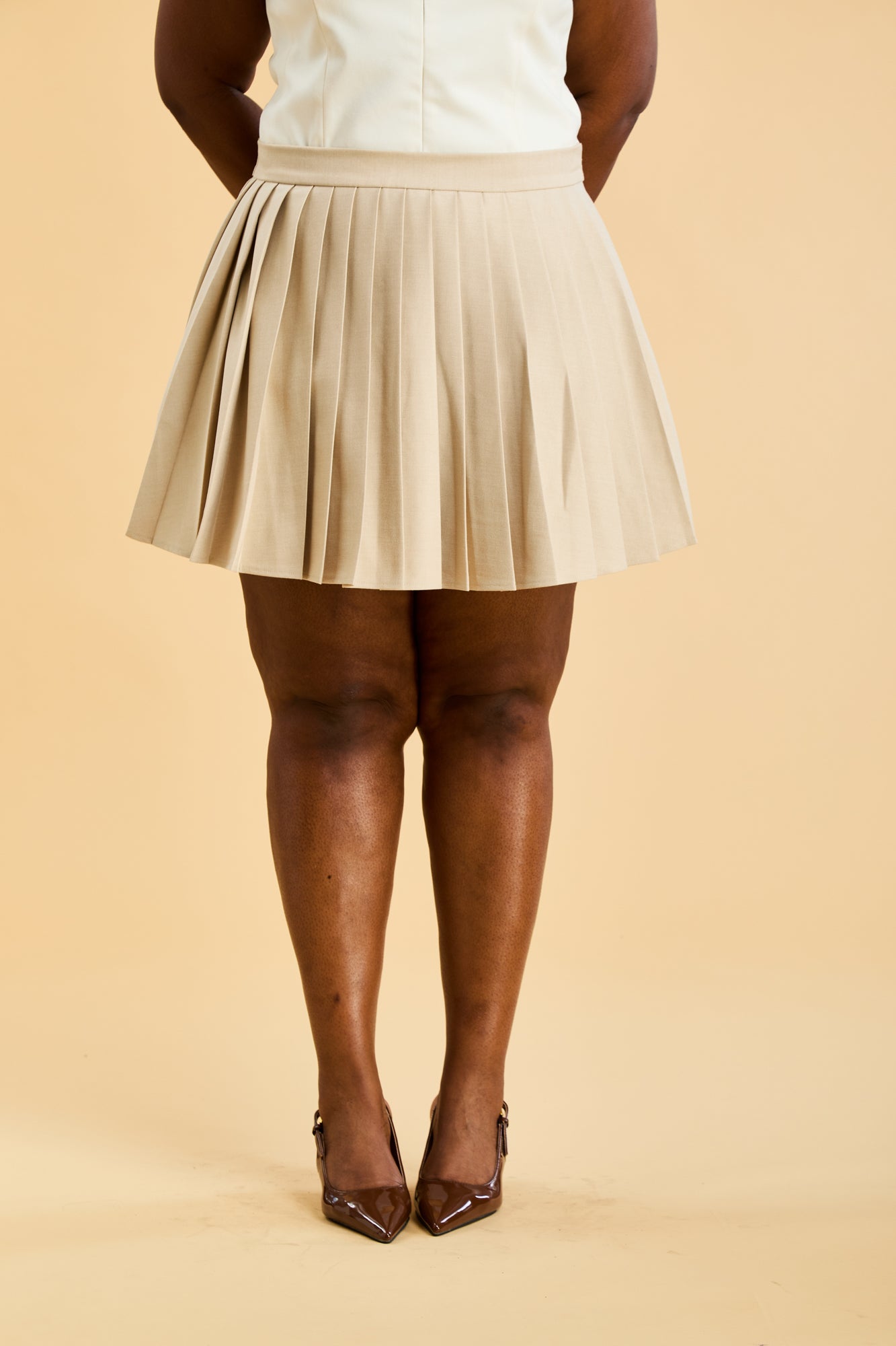 Woman wearing a White colored Triplicity Alice double ended zipper tank top and a Oat Pleated Elisabeth mini skirt on a beige background close up front view