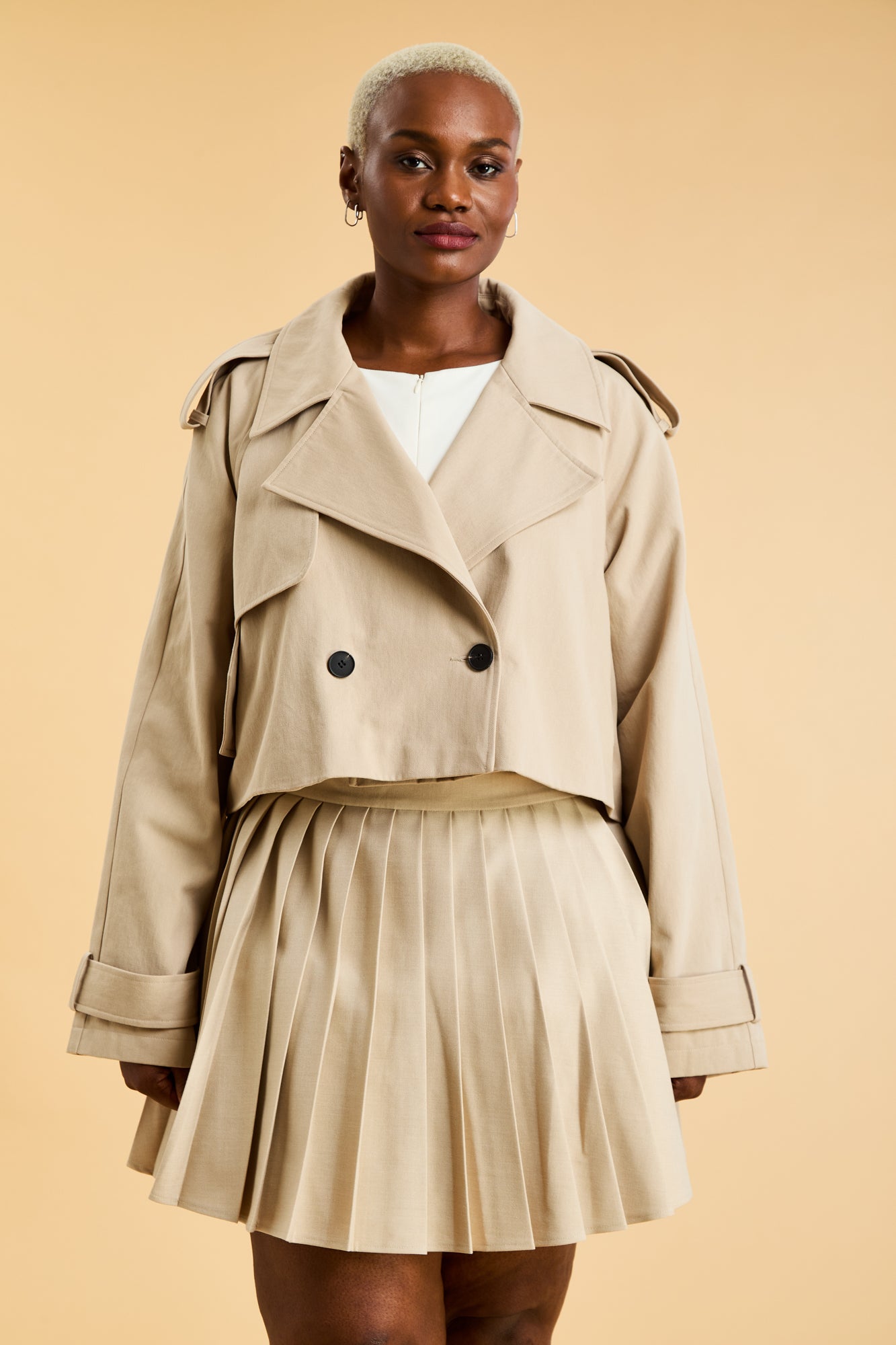 Woman wearing a Triplicity Victoria cropped trench coat and a Oat Pleated Elisabeth mini skirt on a beige background close up front view