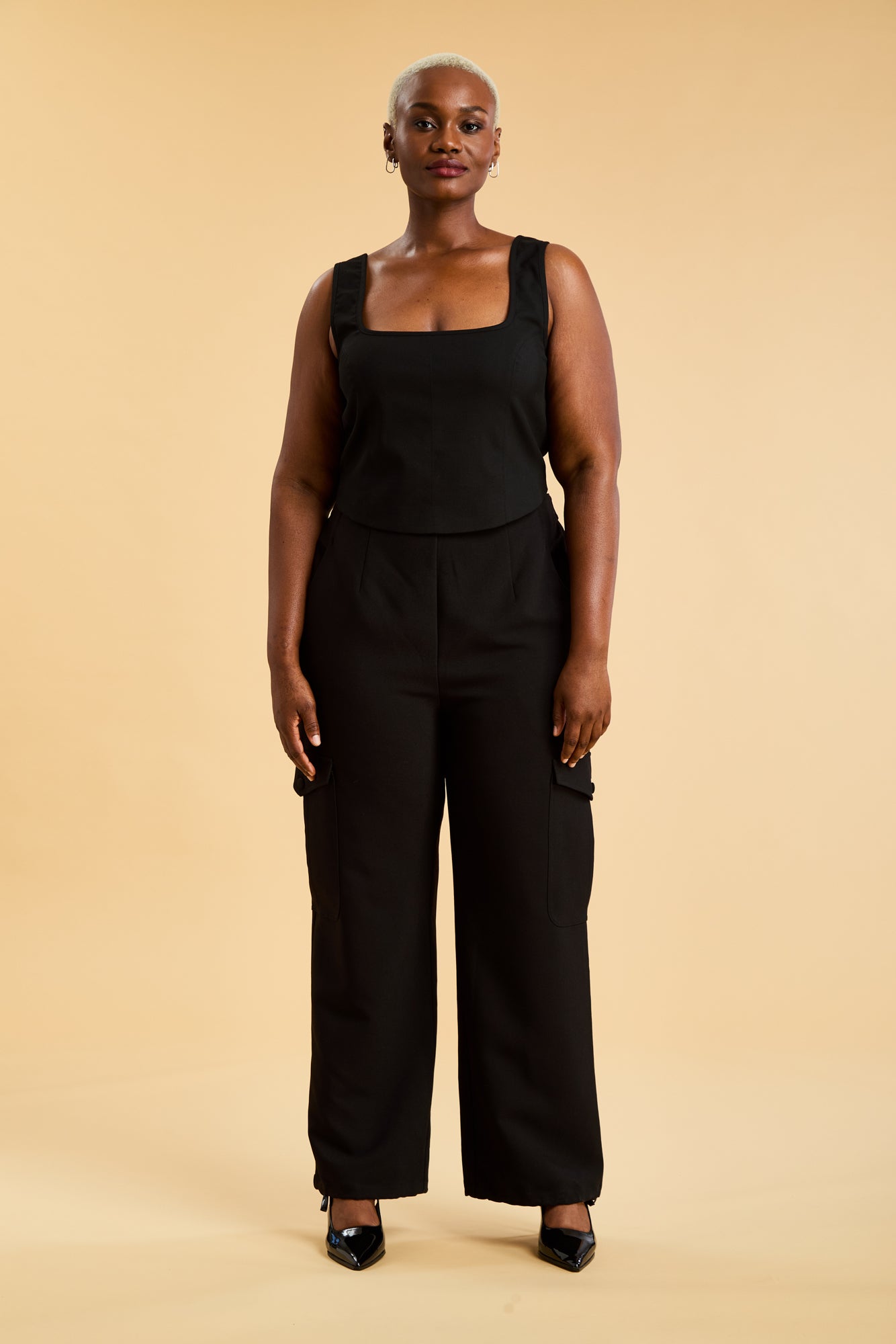 Woman wearing a Black Triplicity sleeveless Eloise tank up against a beige background front view full body