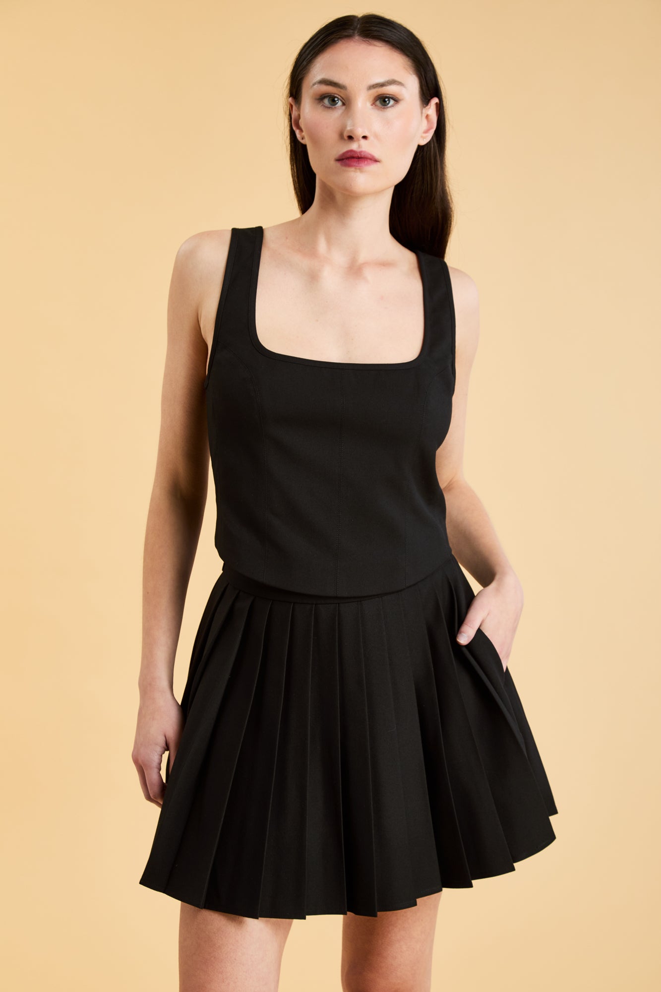 Woman wearing a Triplicity Eloise Fitted Tank Top and a Black Pleated Elisabeth mini skirt on a beige background close up front view