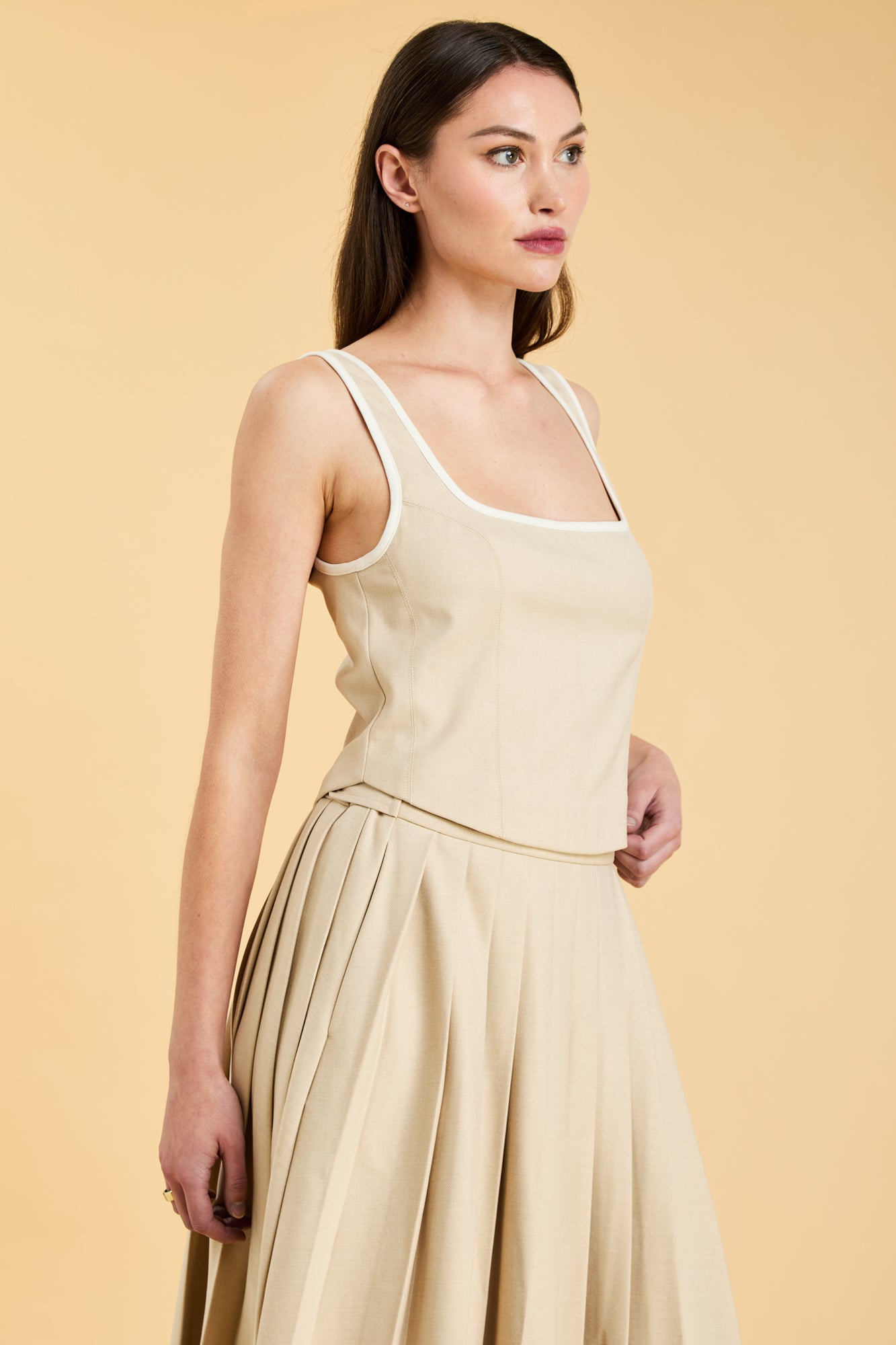 Woman wearing a Oat colored Triplicity Eloise Tank Top on a Oat colored Elisabeth pleated midi skirt against a beige background side view close up