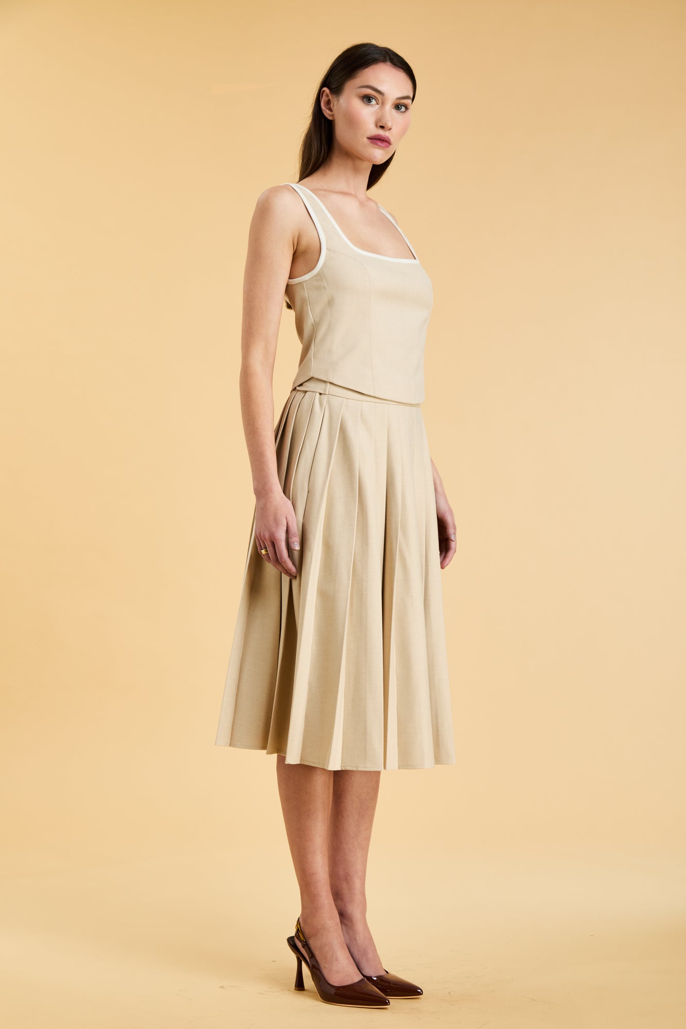 Close up of a woman wearing a Triplicity Oat Elisabeth pleated mini skirt with her leg sticking out of the skirt front slit against a beige background three quarter view