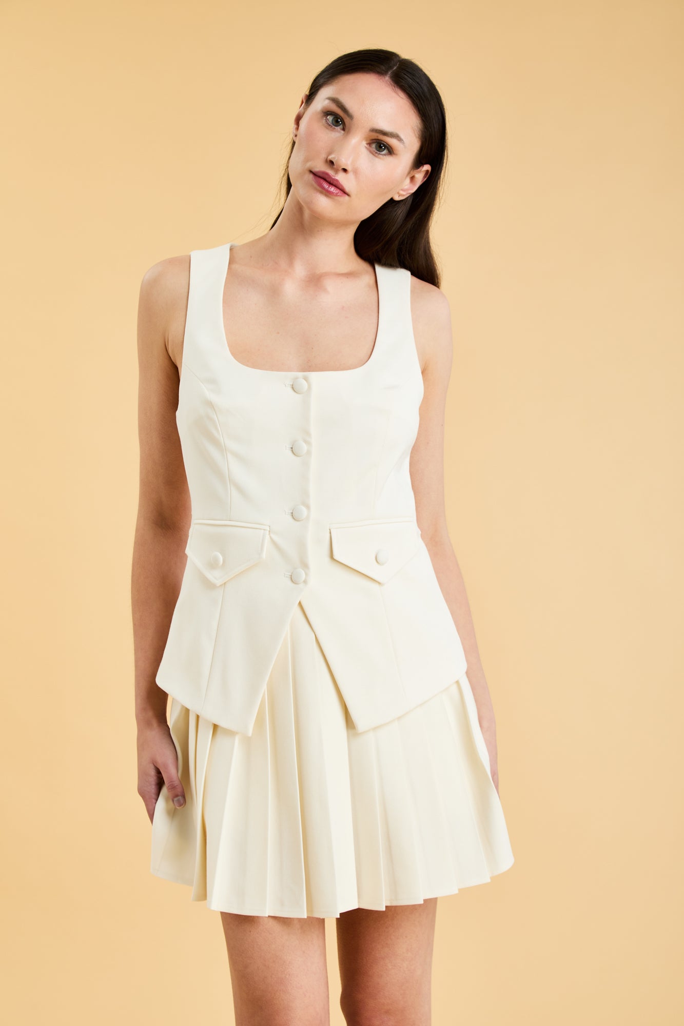 Woman wearing a White Triplicity Georgiana vest top on a Triplicity White pleated midi skirt against a beige background front view close up