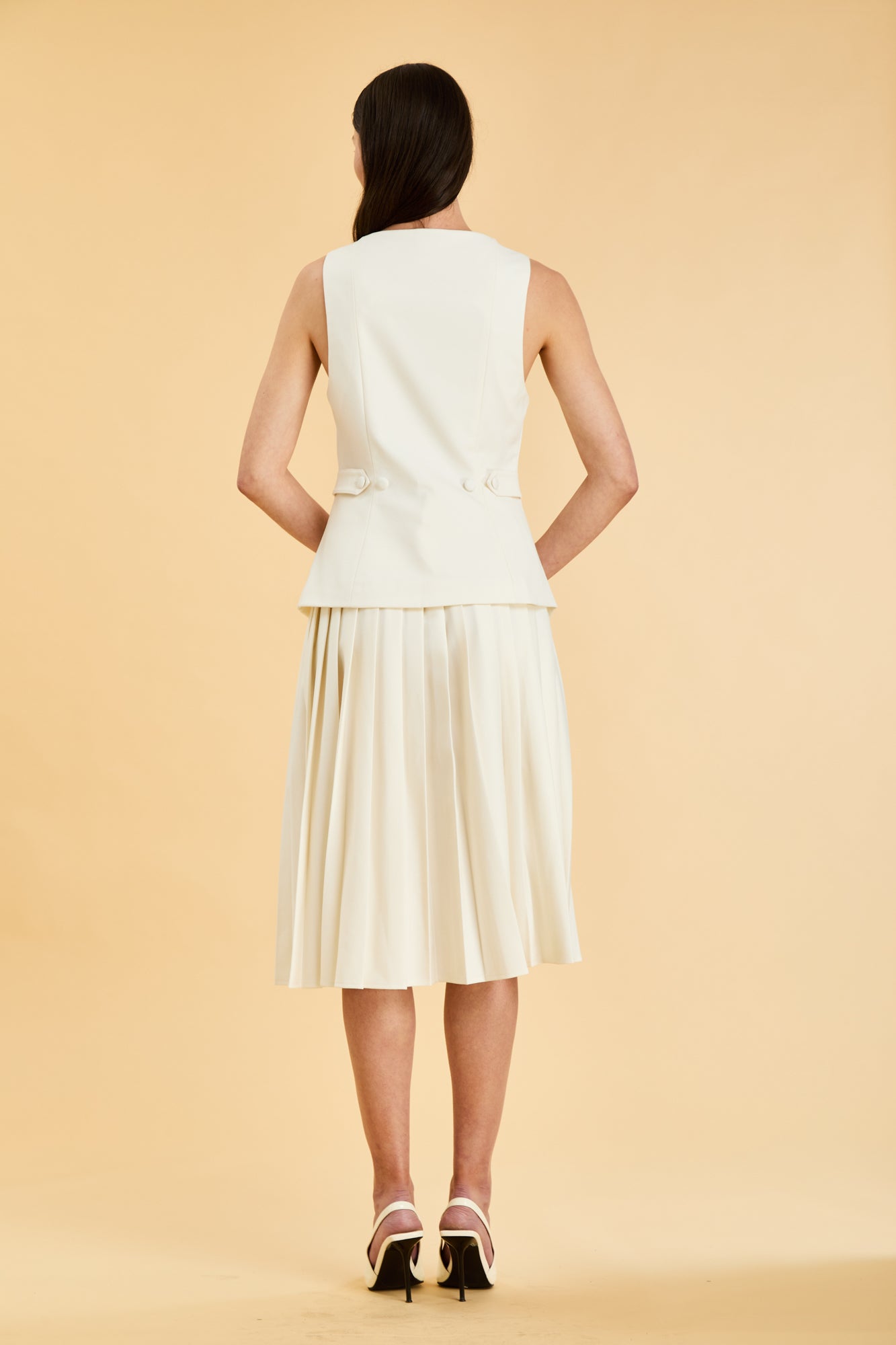 Woman wearing a White Triplicity Georgiana vest top on a Triplicity White pleated midi skirt against a beige background back view full body