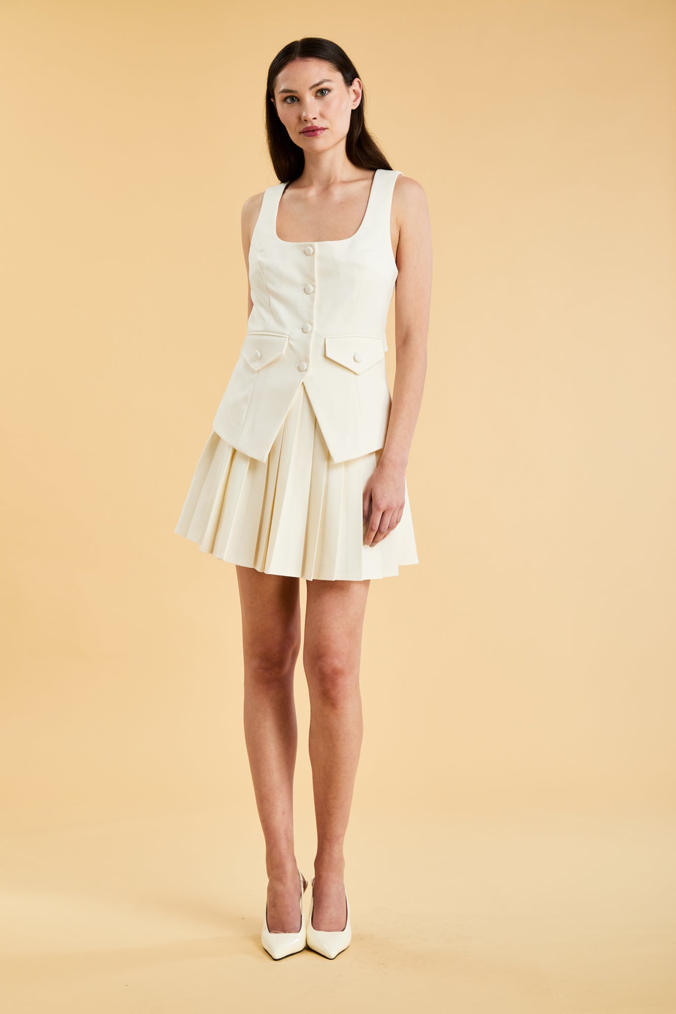 Woman wearing a White Triplicity Georgiana vest top on a Triplicity White pleated min skirt against a beige background front view full body
