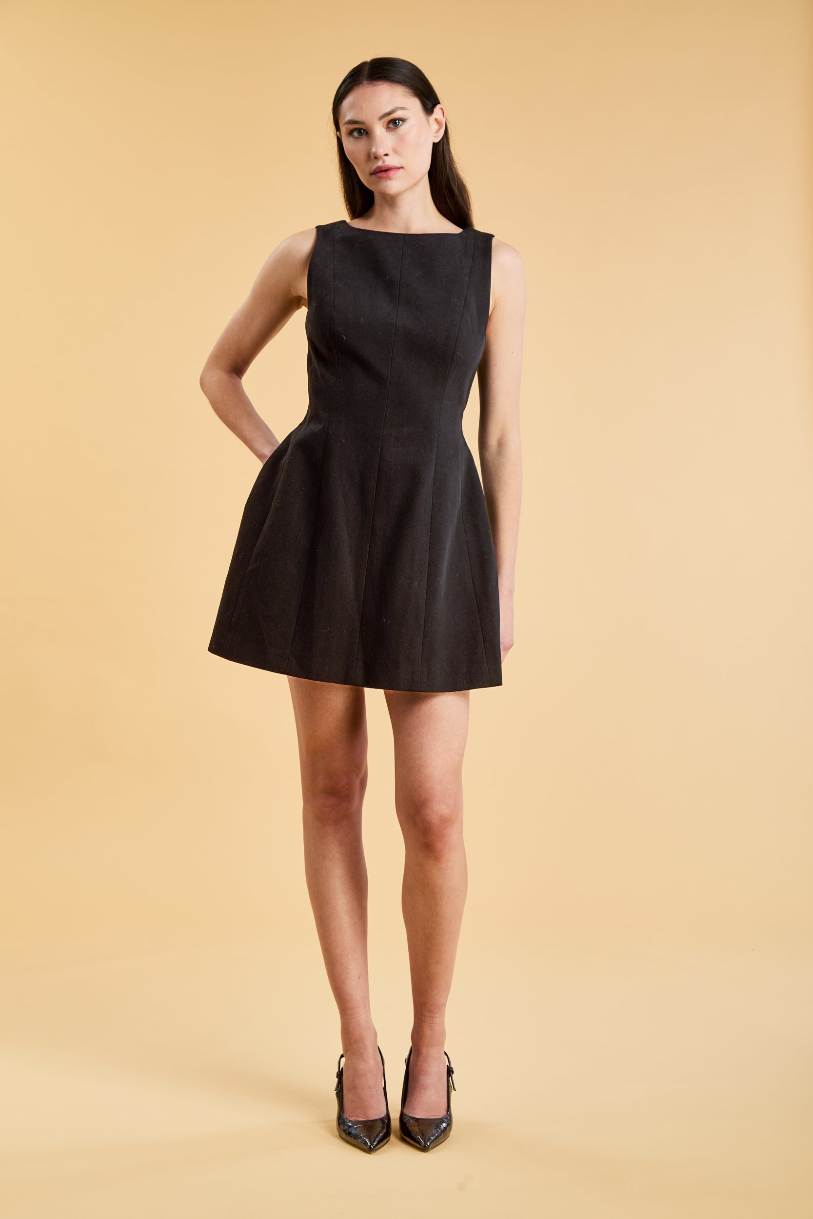 Full body view of a woman wearing the Triplicity Louise Sleeveless Mini Dress on a beige background