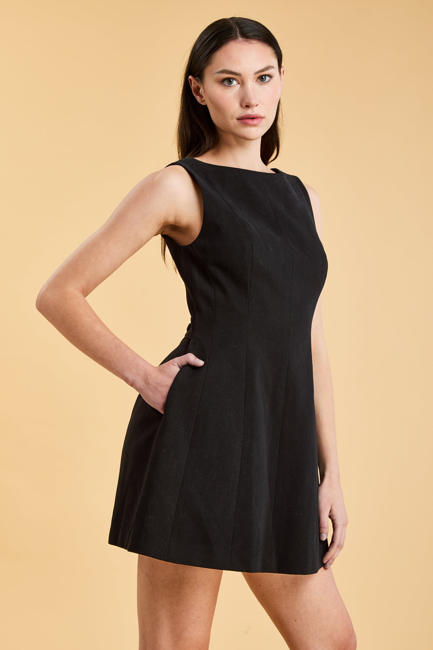 Close up view of a woman wearing the Triplicity Louise Sleeveless Mini Dress on a beige background