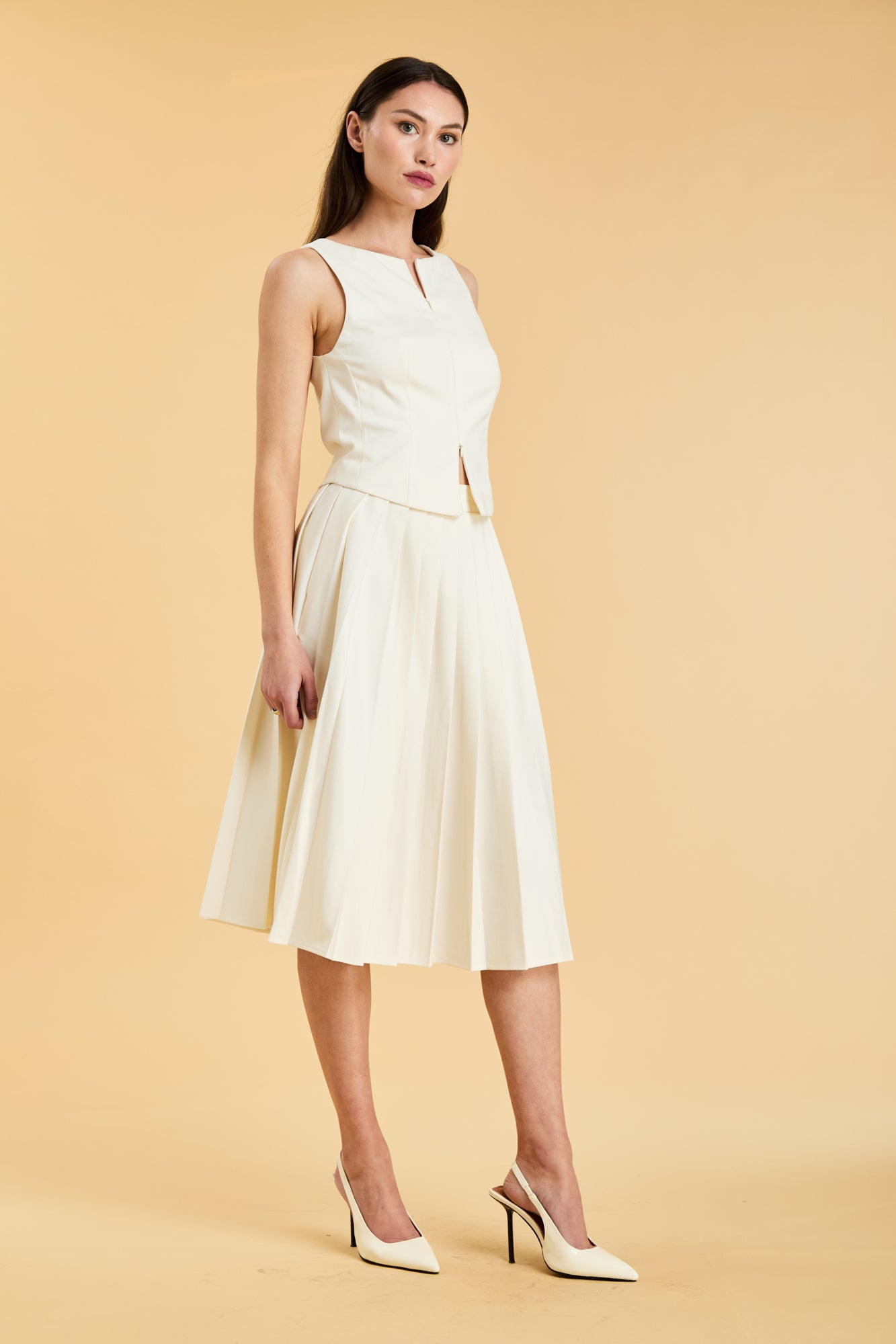 Woman wearing a White colored Triplicity double ended zipper Alice top and White Triplicity pleated midi skirt against a beige background three quarter view full body
