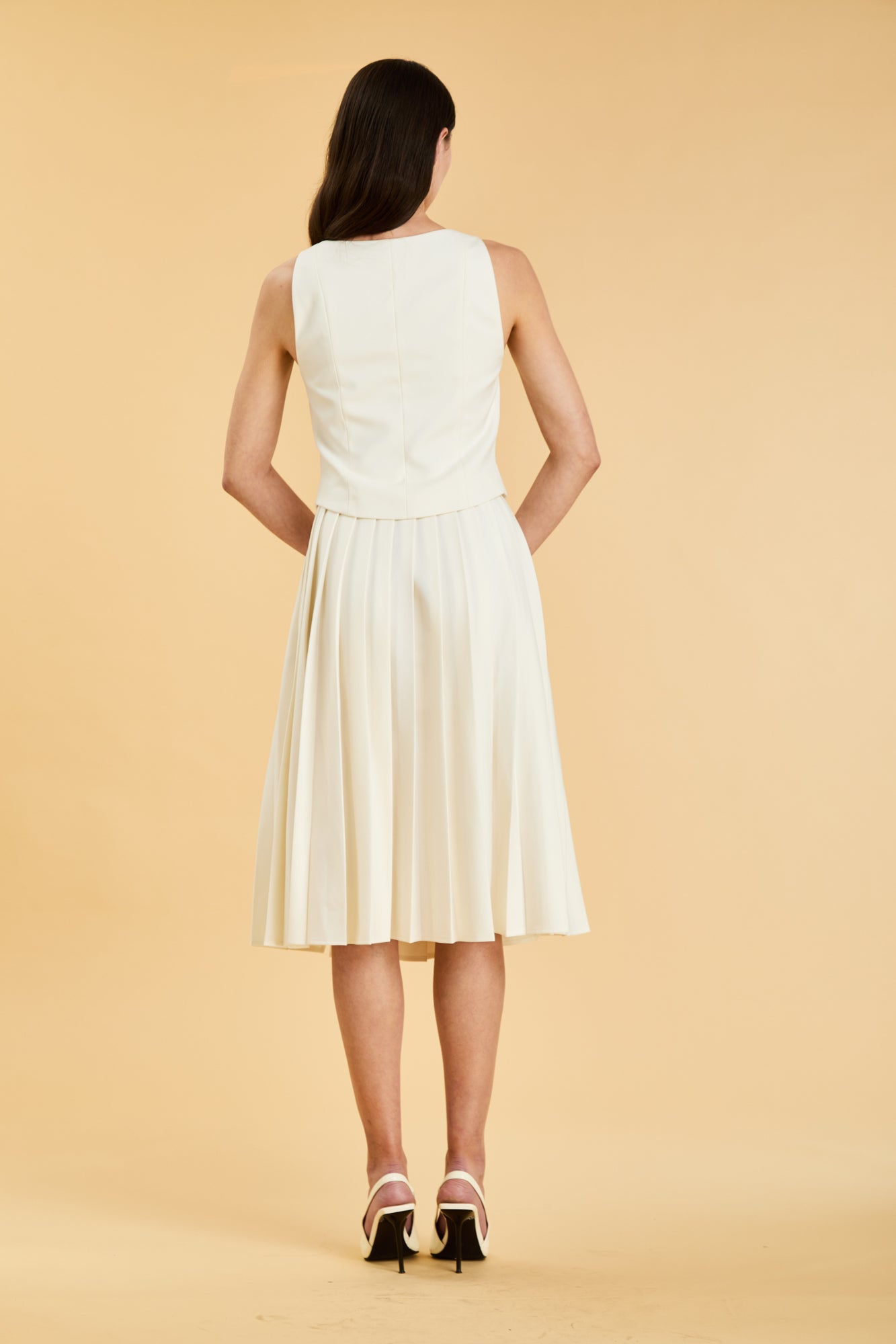Woman wearing a White colored Triplicity double ended zipper Alice top and White Triplicity pleated midi skirt against a beige background back view full body