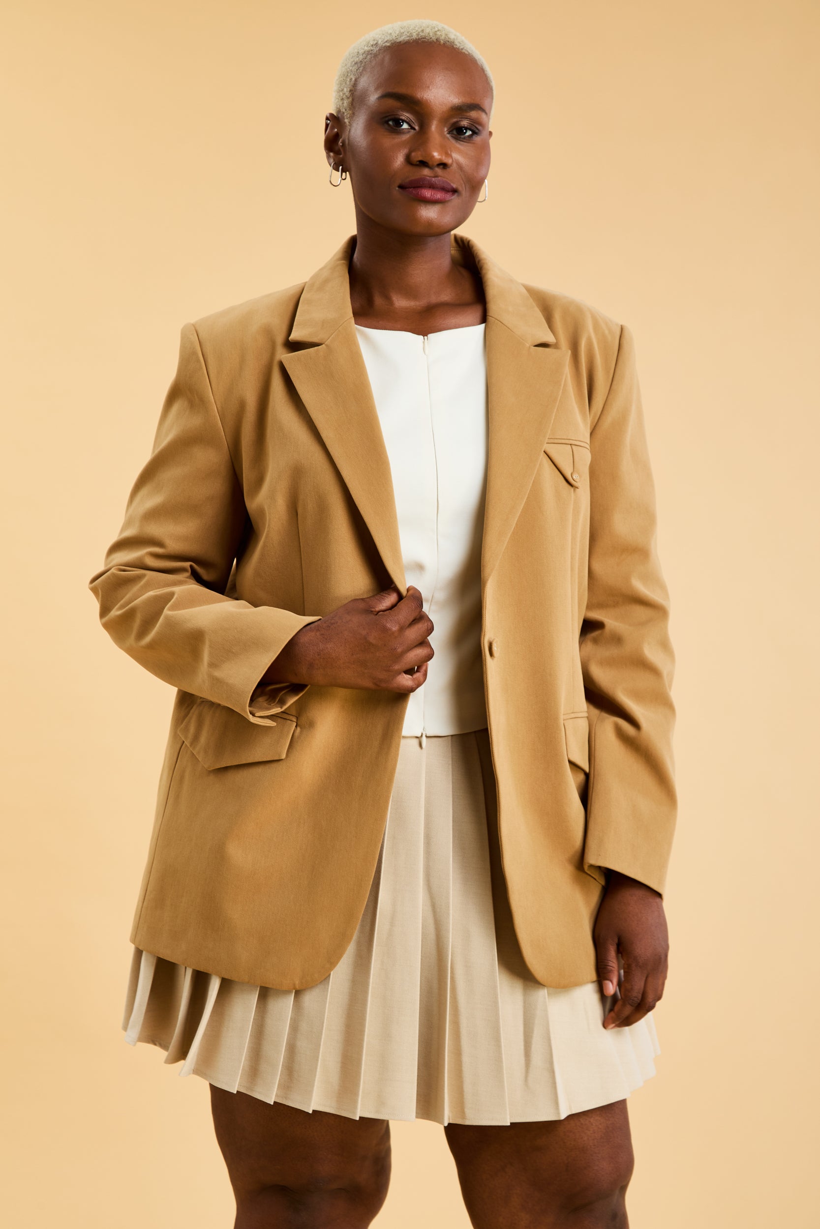 Woman wearing a tan Camel colored blazer over a beige top and skirt on a beige background