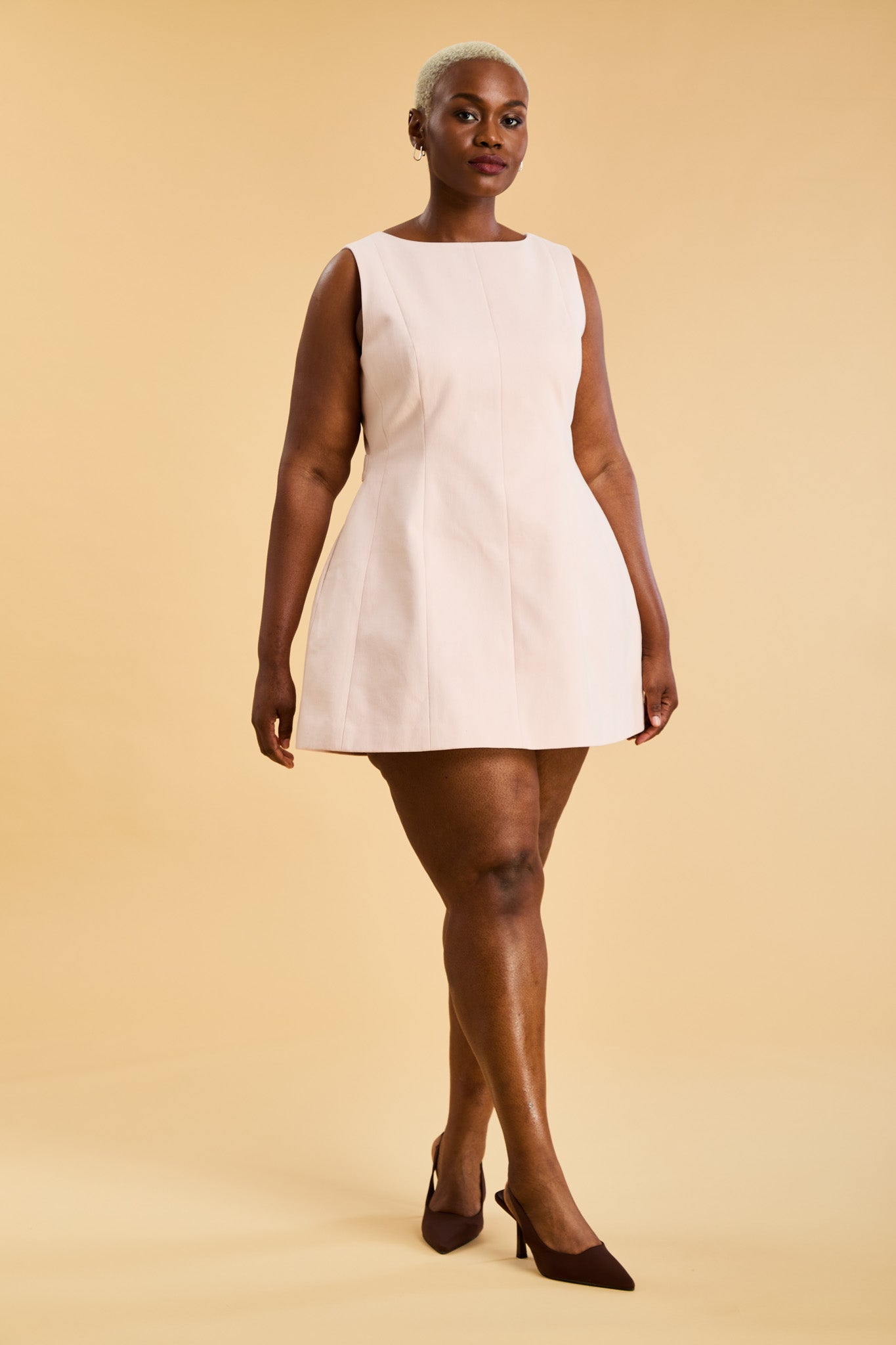 Woman wearing the Triplicity blush pink colored Louise mini sleeveless dress on a beige background
