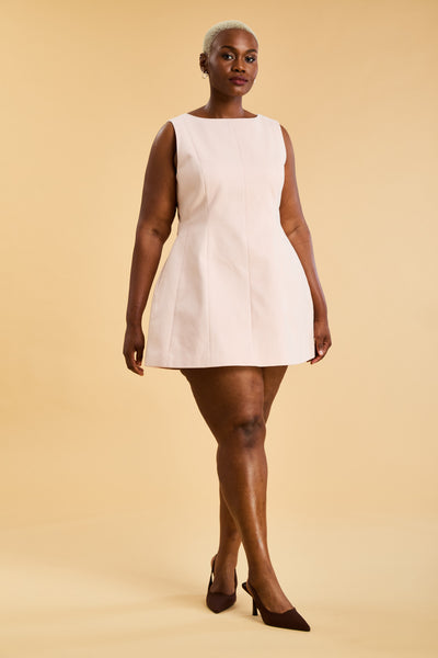 Woman wearing the Triplicity blush pink colored Louise mini sleeveless dress on a beige background