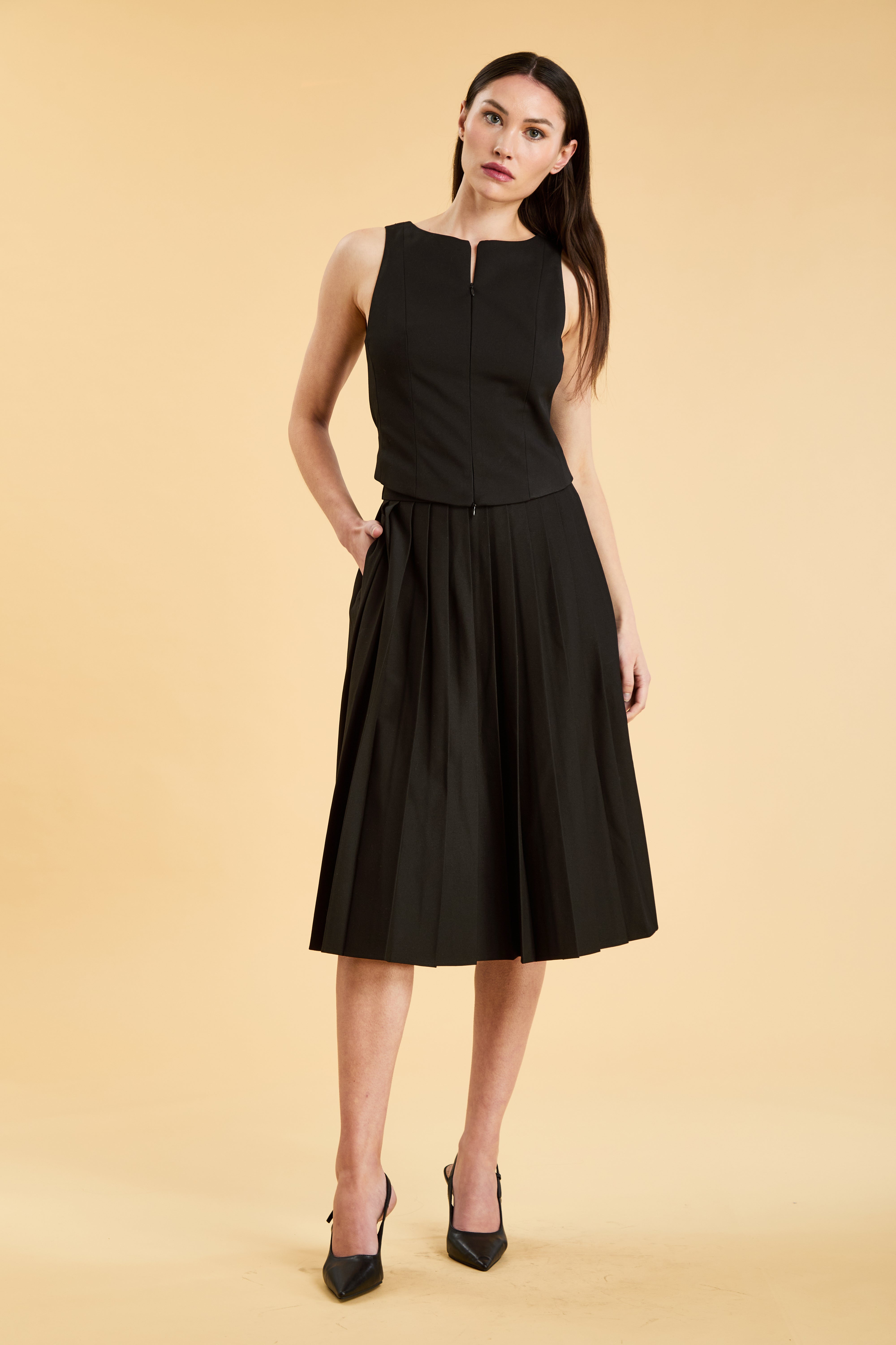 Person wearing a Black Alice double ended zipper Top on a Black pleated Elisabeth Mini skirt against a beige background