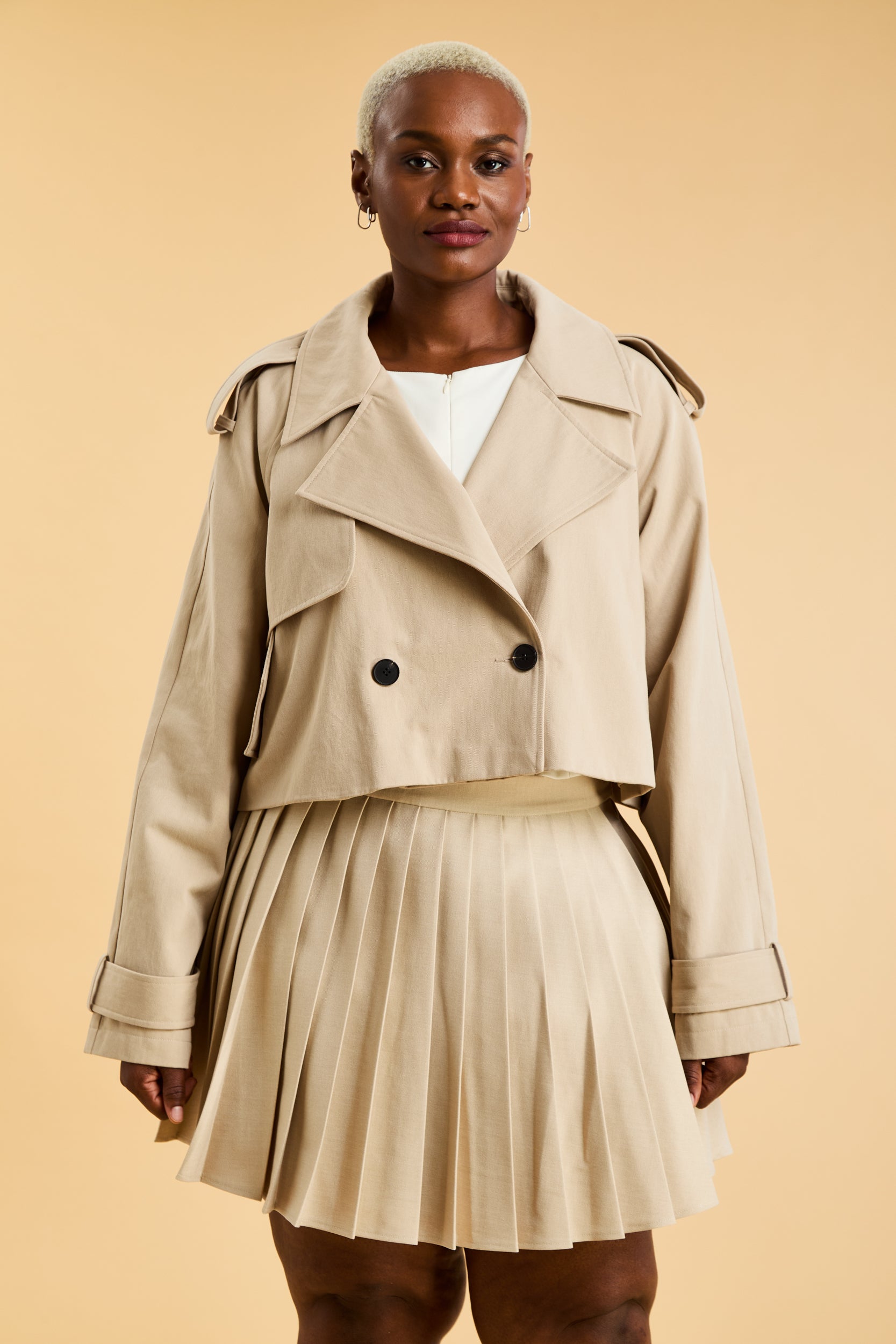 Woman wearing a beige cropped trench coat jacket in stone color against a beige background
