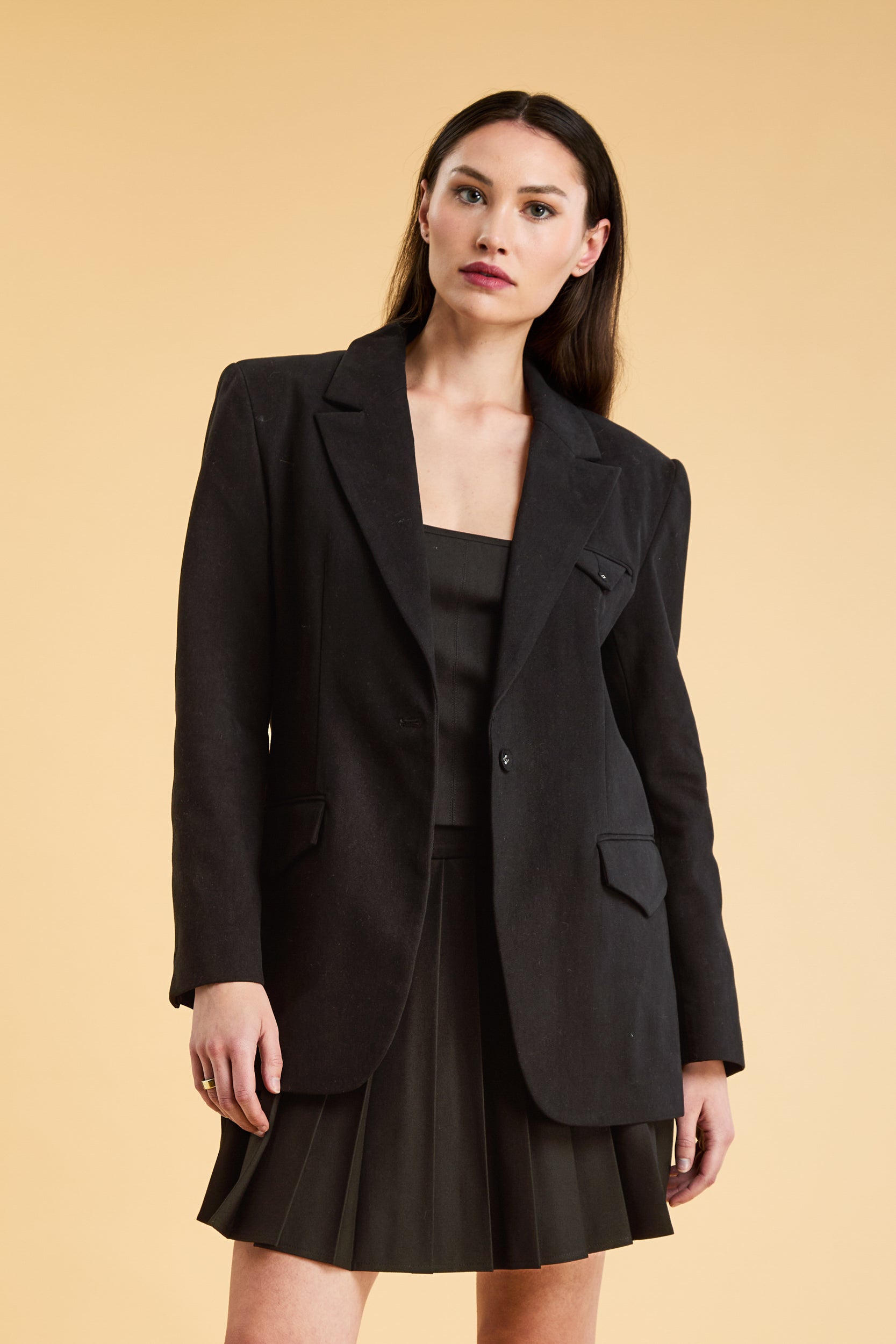 Woman wearing a Black oversized tailored Triplicity blazer over a black pleated skirt on a beige background close up